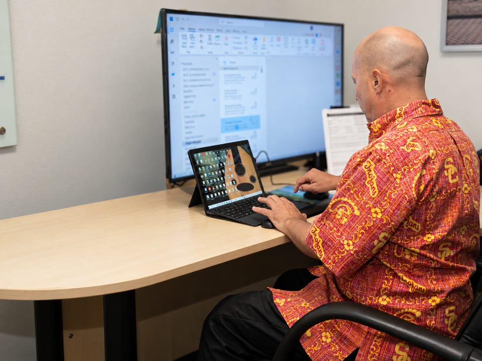 A man in a red patterned shirt types on a tablet keyboard at a desk facing a large monitor in an office