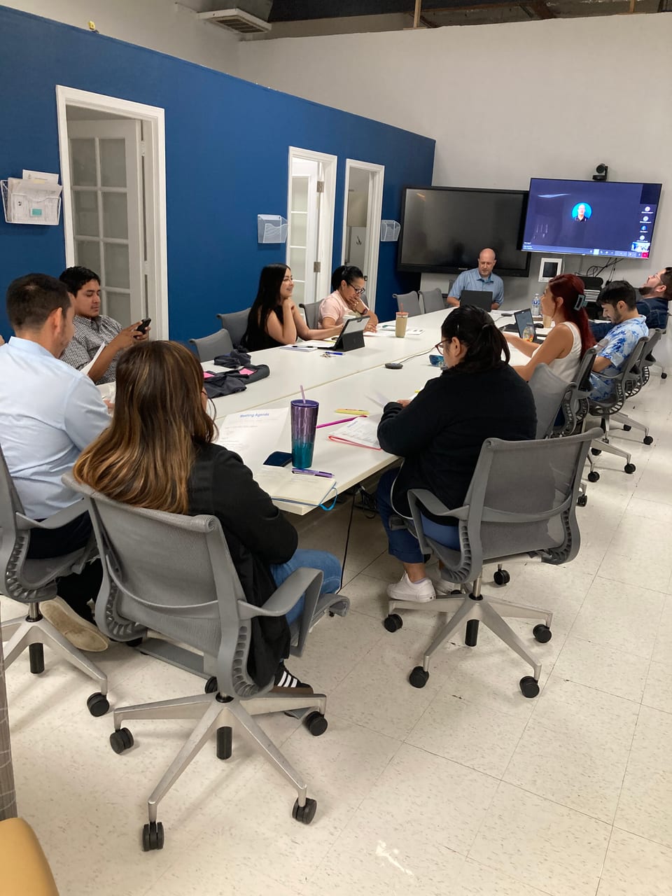 Ten colleagues sit around a long white table in a meeting room with a blue wall, one more joining on the wall screen