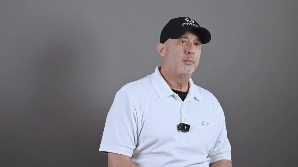 Pete Velez seated in a black UFG cap and white polo, looking aside, a frame from the film Coaches, Not Supervisors